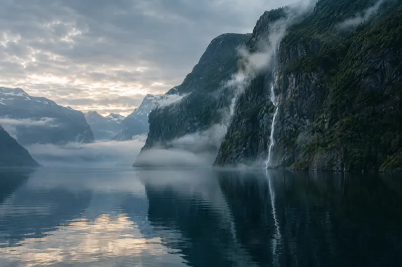 Norwegischer Fjord am Morgen mit steilen Felswänden, Nebel über dem Wasser und einem Wasserfall